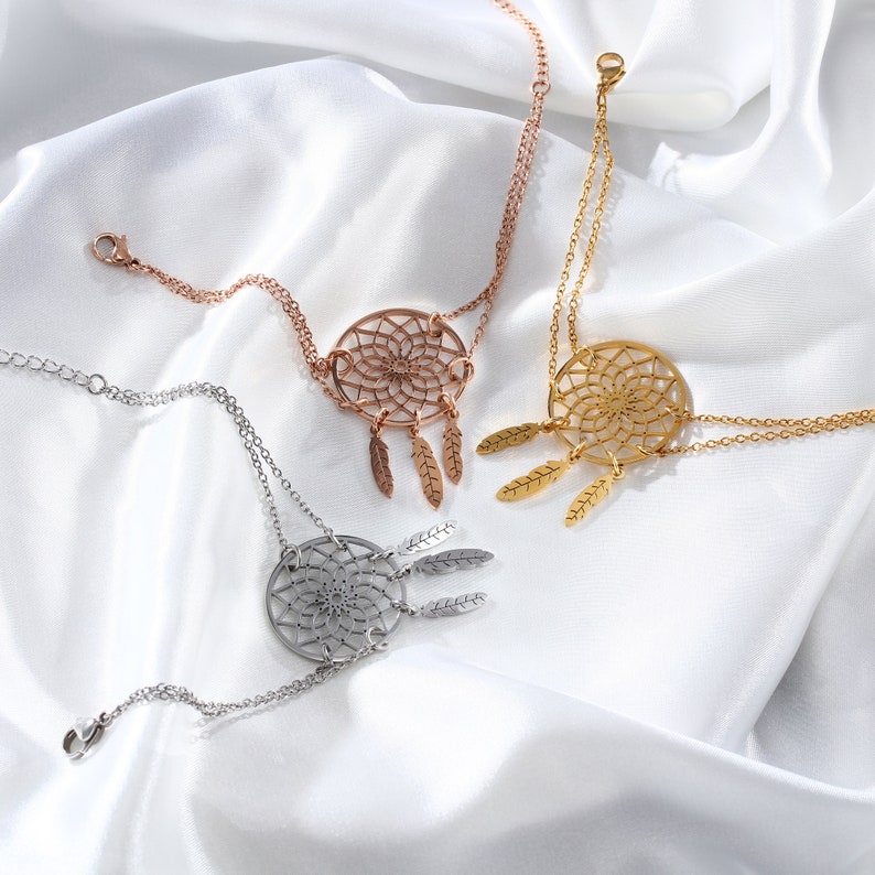 May include: Three gold-toned dreamcatcher bracelets with delicate feather charms. The bracelets are layered on a white fabric background.