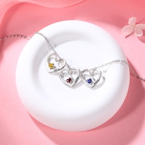 May include: A silver necklace with three heart-shaped pendants, each containing a gemstone. The pendants are engraved with the names "Daniel", "Ashley", and "Jessica". The gemstones are yellow, red, and blue.