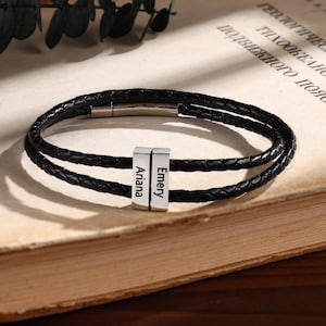 Puede incluir: Pulsera de cuero trenzado negro con dos hebras. La pulsera presenta dos cuentas rectangulares plateadas con los nombres "Ariana" y "Emery" grabados. La pulsera está sobre un libro.