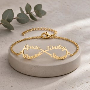 May include: A gold-toned metal bracelet featuring an infinity symbol centerpiece. The infinity loop is integrated with four names written in a cursive script: Gracie, Kinsley, Dominic, and Steven. The bracelet includes a delicate chain and a lobster clasp.