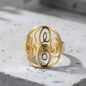 Pode incluir: Um anel dourado com um design de monograma com as letras B, E e A num padrão circular vazado. O anel é feito de metal polido e brilhante e assenta numa superfície clara. O anel é uma joia.
