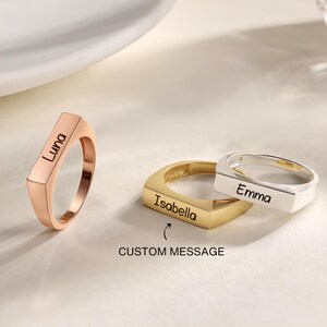 Pode incluir: Três anéis personalizados em ouro rosa, ouro e prata. Os anéis têm um design retangular com os nomes gravados: "Luna", "Isabella" e "Emma". A imagem também inclui o texto "CUSTOM MESSAGE".