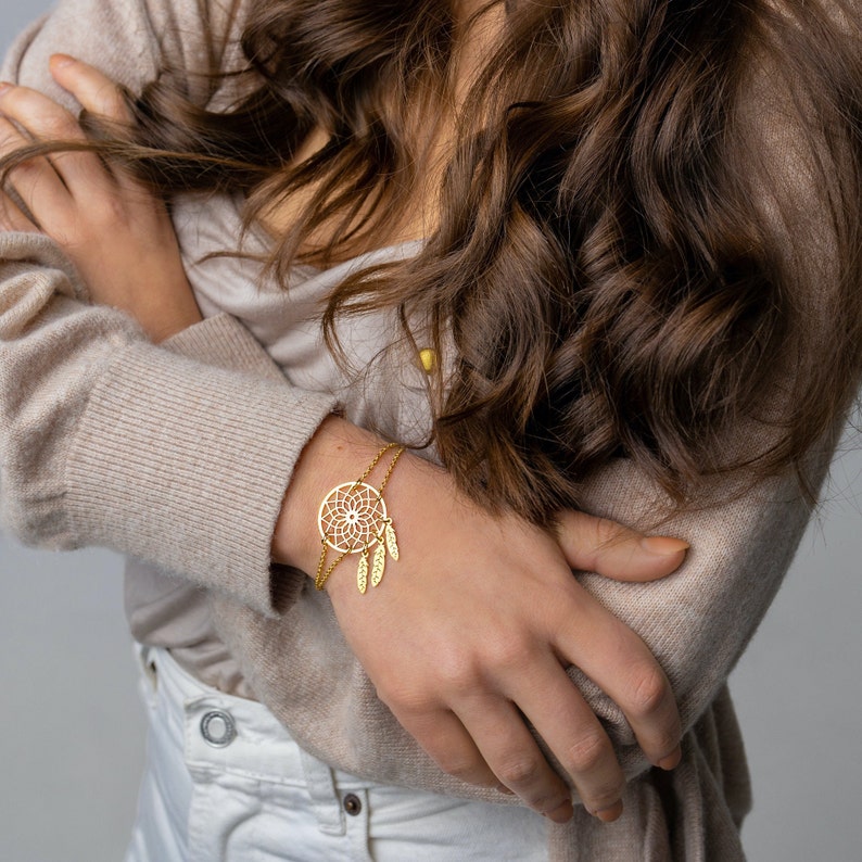 May include: A gold-toned bracelet with a dreamcatcher charm and dangling feathers. The bracelet is on a woman's wrist.