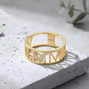 Pode incluir: Um anel dourado com algarismos romanos recortados ao redor da banda. O anel está sobre uma superfície texturizada cinza claro. O design é simples e elegante, com uma estética moderna. O diâmetro do anel é de aproximadamente 2 cm.