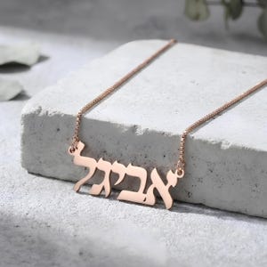 May include: A rose gold necklace featuring a Hebrew name pendant. The pendant is attached to a fine chain. The necklace is presented on a white, textured surface.