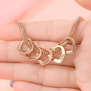 Puede incluir: Un collar dorado con una cadena y cinco colgantes en forma de corazón. Cada colgante en forma de corazón tiene un nombre grabado: Brodie, Hanna, Lainey, Tyler y Oliver. El collar se muestra en una mano.