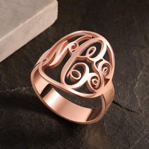 May include: A rose gold ring featuring a large, circular face with an intricate, openwork monogram design. The letters are interconnected in a flowing, cursive script style, creating a decorative centerpiece that sits atop a smooth, polished band.