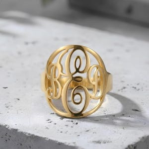 May include: A gold-colored ring featuring a monogram design with the letters B, E, and A in a circular, openwork pattern. The ring is made of a shiny, polished metal and sits on a light-colored surface. The ring is a piece of jewelry.