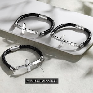Può includere: Tre braccialetti in pelle intrecciata nera con ciondoli a forma di croce argentata. Ogni croce ha un'iscrizione personalizzata: "Alexander", "Charlotte❤Richard" e "JOHN 14:6". I braccialetti sono esposti su una superficie bianca.