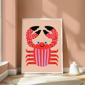 May include: A framed wall art print featuring a stylized red crab illustration on a light peach background. The crab has black-tipped claws with red spots, a red body with vertical light purple stripes, and two small blue eyes with a simple smile.