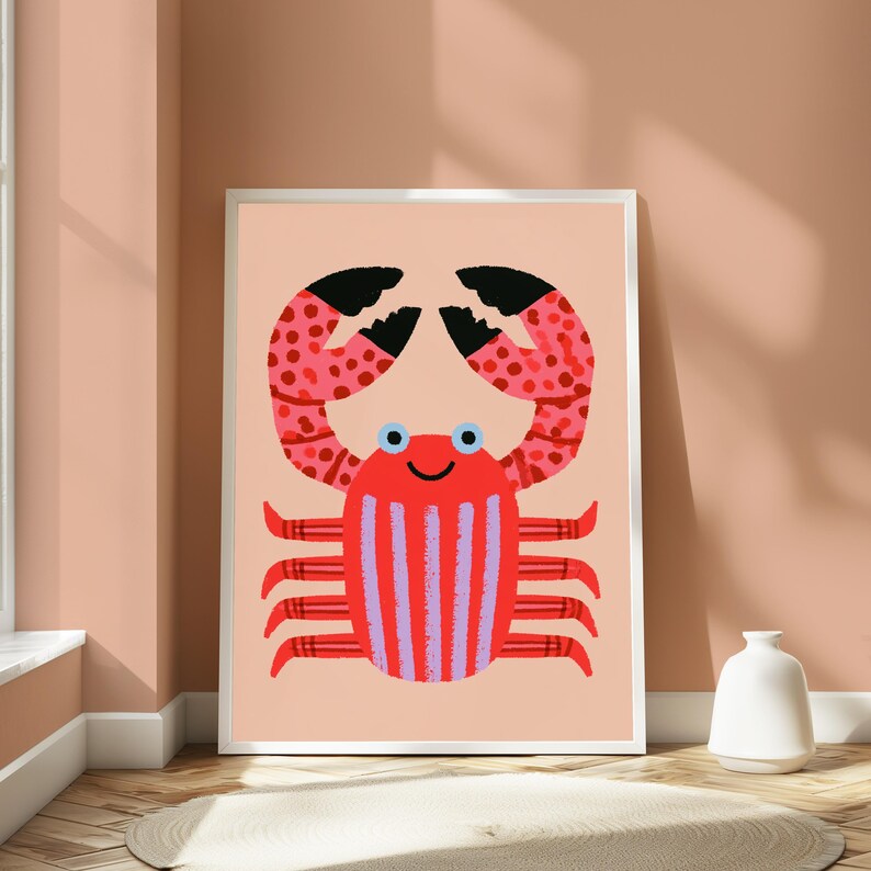 May include: A framed wall art print featuring a stylized red crab illustration on a light peach background. The crab has black-tipped claws with red spots, a red body with vertical light purple stripes, and two small blue eyes with a simple smile.