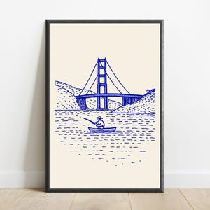 May include: A framed wall art print featuring a blue ink line drawing on a cream background. The illustration depicts a person in a small boat fishing on the water with a large suspension bridge and hilly terrain visible in the background.