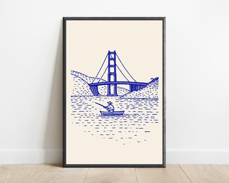 May include: A framed wall art print featuring a blue ink line drawing on a cream background. The illustration depicts a person in a small boat fishing on the water with a large suspension bridge and hilly terrain visible in the background.
