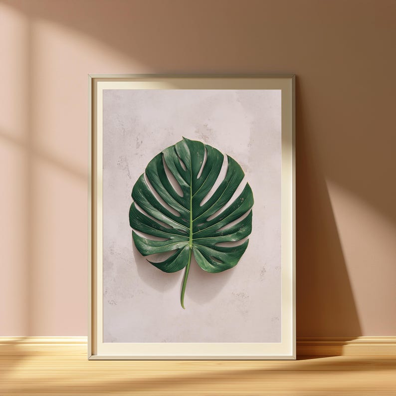 May include: A framed wall art print featuring a single, large, dark green monstera leaf centered against a light, textured, off-white background. The artwork is enclosed in a thin, metallic frame, presenting a clean and focused botanical composition.