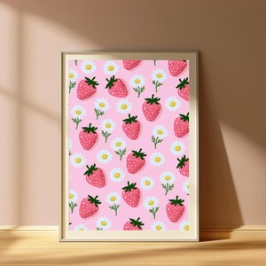 May include: A framed wall art print featuring a repeating pattern of red strawberries with green stems and white daisies with yellow centres on a solid light pink background. The frame is a simple, light-coloured wood with a thin, clean border.