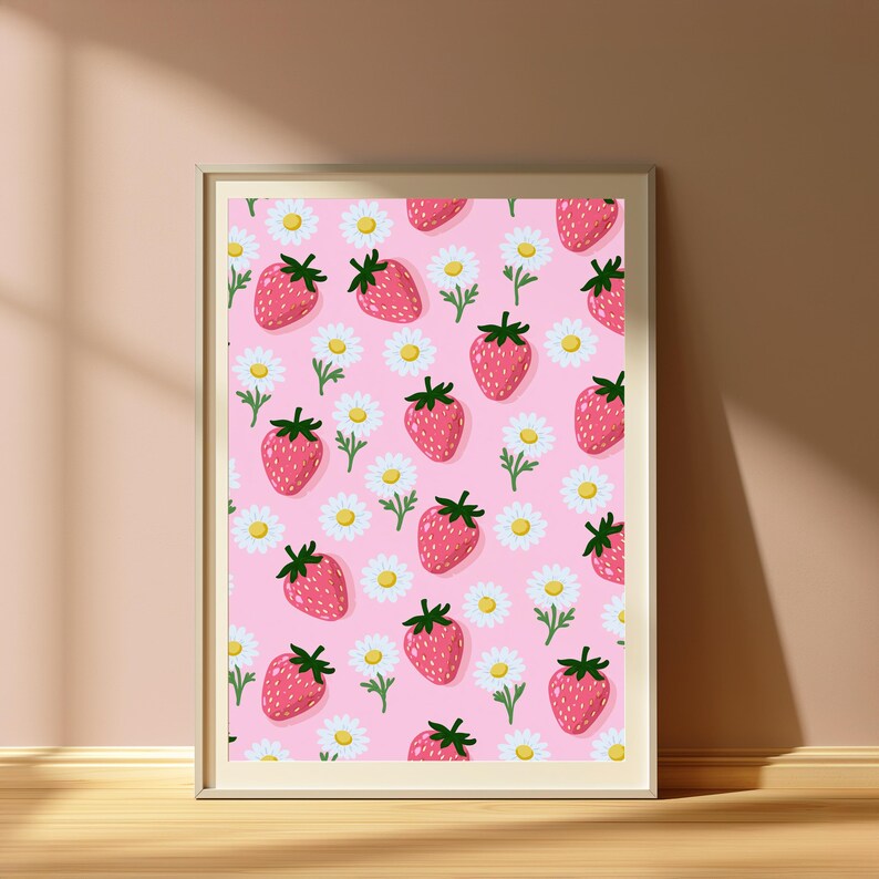May include: A framed wall art print featuring a repeating pattern of red strawberries with green stems and white daisies with yellow centres on a solid light pink background. The frame is a simple, light-coloured wood with a thin, clean border.