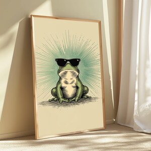 May include: A framed wall art print featuring a green frog wearing black sunglasses. The frog is centered against a light cream background with a radiating teal sunburst pattern behind it. The illustration uses detailed line work and shading.