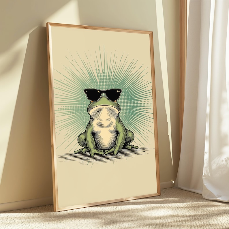 May include: A framed wall art print featuring a green frog wearing black sunglasses. The frog is centered against a light cream background with a radiating teal sunburst pattern behind it. The illustration uses detailed line work and shading.