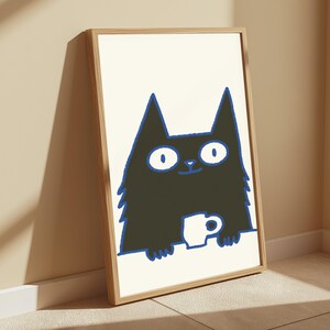 May include: A framed wall art print featuring a black cat illustration with wide white eyes and a blue outline. The cat is positioned behind a white surface, holding a small white mug. The artwork is enclosed in a light-colored wooden frame.