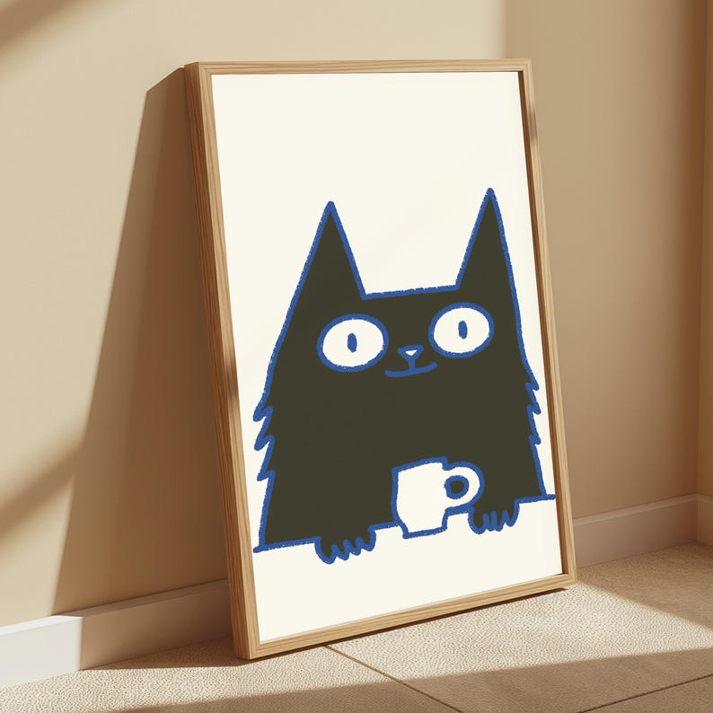 May include: A framed wall art print featuring a black cat illustration with wide white eyes and a blue outline. The cat is positioned behind a white surface, holding a small white mug. The artwork is enclosed in a light-colored wooden frame.