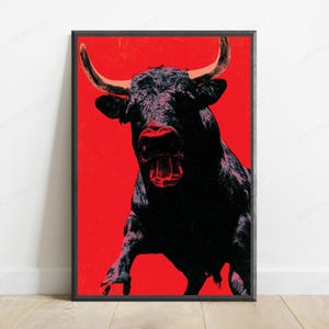 K&ouml;nnte beinhalten: Ein gerahmter vertikaler Wandkunstdruck mit einer kontrastreichen Illustration eines schwarzen Stiers mit hellen H&ouml;rnern vor einem einfarbigen, leuchtend roten Hintergrund. Der Stier ist in einer frontalen Haltung mit offenem Maul dargestellt.