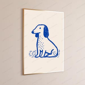 May include: A framed wall art print featuring a blue line drawing of a sitting dog on a cream-colored background. The illustration uses a textured, crayon-like stroke to depict the canine figure, which is centered within a thin gold metal frame.