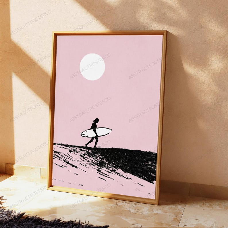 May include: A rectangular framed wall art print featuring a light pink background with a white circular sun. A black silhouette of a person carrying a surfboard walks across a textured black landscape, creating a high-contrast graphic composition.