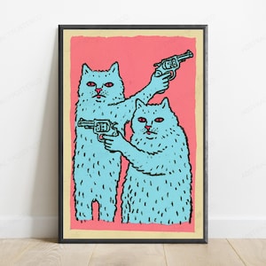 May include: A framed wall art print featuring two light blue cats with red eyes against a solid pink background. Each cat is holding a dark blue revolver. The illustration uses bold black outlines and is enclosed in a simple black frame.