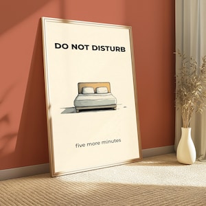Puede incluir: Un p&oacute;ster enmarcado con las palabras "DO NOT DISTURB" en la parte superior y "five more minutes" en la inferior. El p&oacute;ster presenta una ilustraci&oacute;n de una cama con dos almohadas. El p&oacute;ster est&aacute; apoyado contra una pared roja.