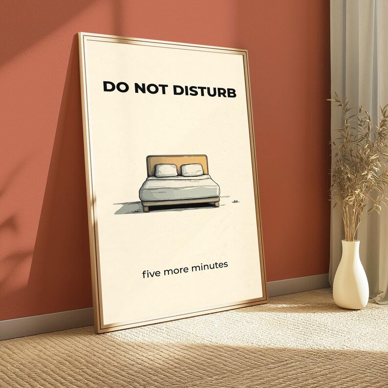 Puede incluir: Un p&oacute;ster enmarcado con las palabras "DO NOT DISTURB" en la parte superior y "five more minutes" en la inferior. El p&oacute;ster presenta una ilustraci&oacute;n de una cama con dos almohadas. El p&oacute;ster est&aacute; apoyado contra una pared roja.