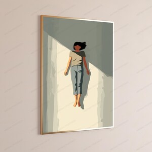 May include: A framed wall art print featuring a stylized illustration of a person with dark hair, wearing a beige short-sleeved shirt and blue pants, lying flat on a light-colored surface with sharp geometric shadows cast across the scene.