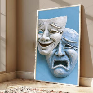 May include: A framed wall art print featuring two theatrical masks against a solid light blue background. One white mask displays a smiling expression, while the overlapping blue mask below it displays a sad, weeping expression with deep-set eyes.