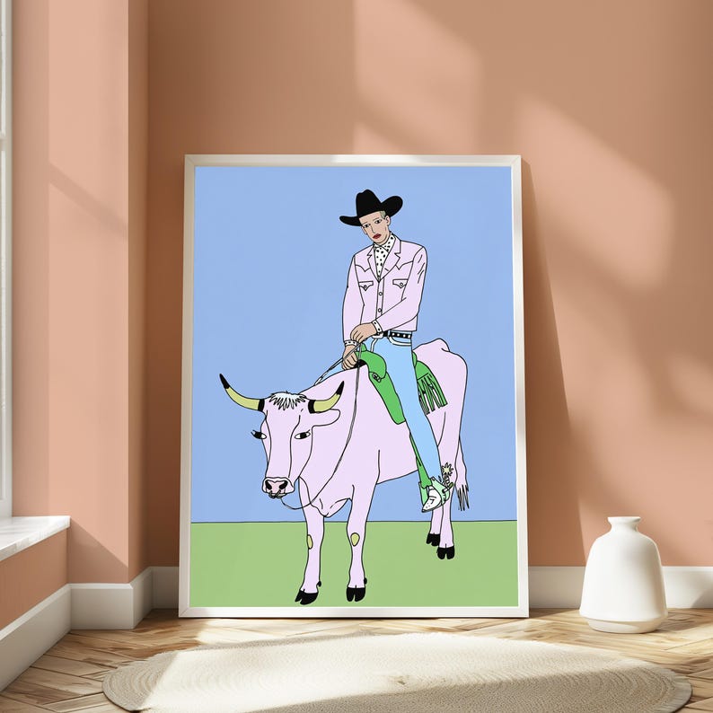 May include: A framed art print featuring a line drawing of a person in a black cowboy hat, pink shirt, and blue jeans riding a pink cow. The background is a light blue sky over a green field. The illustration uses bold black outlines.