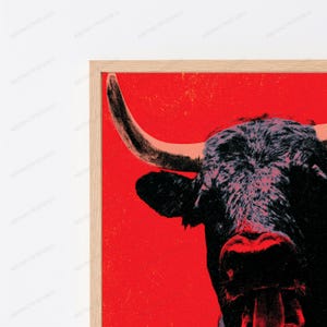 K&ouml;nnte beinhalten: Ein gerahmter Wandkunstdruck mit einem kontrastreichen, stilisierten schwarzen Bullenkopf vor einem leuchtend roten Hintergrund. Der Stier ist im Profil mit einem markanten, geschwungenen Horn und strukturiertem dunklen Fell in einem hellen Holzrahmen dargestellt.