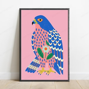 May include: A framed wall art print featuring a stylized falcon illustration on a solid pink background. The bird has a blue head and wings with yellow accents, a pink chest with blue spots, and a central white flower with green leaves.