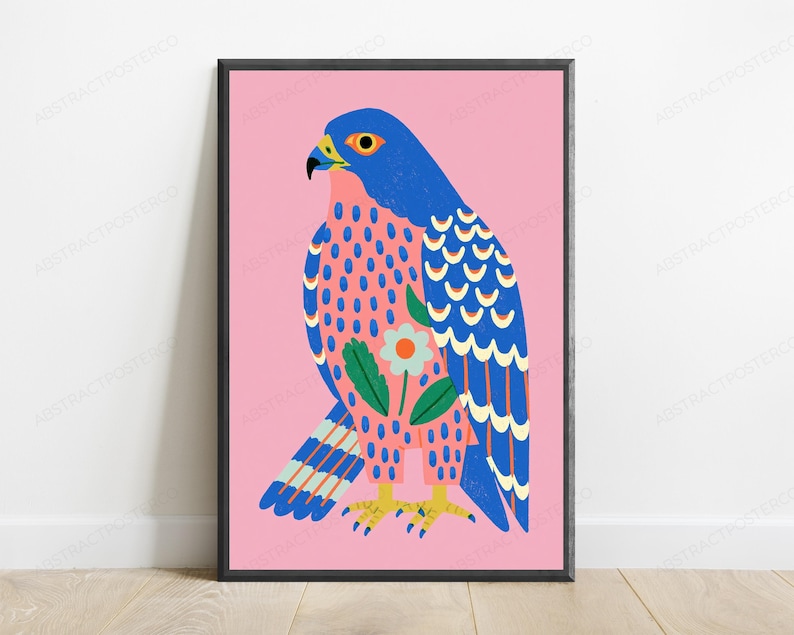 May include: A framed wall art print featuring a stylized falcon illustration on a solid pink background. The bird has a blue head and wings with yellow accents, a pink chest with blue spots, and a central white flower with green leaves.