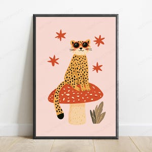 Op de afbeelding: Een ingelijste wandkunstprint met een gele luipaard met zwarte vlekken die een ronde oranje zonnebril draagt, zittend op een grote rode paddenstoel met witte stippen. De achtergrond is lichtroze met vier rode sterren en een kleine groene plant.
