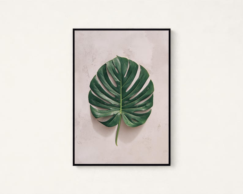 May include: A framed wall art print featuring a single, large, dark green monstera leaf with characteristic splits and holes, centered against a light, textured, off-white background. The artwork is enclosed in a thin, black rectangular frame.