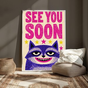 May include: A rectangular wall poster featuring the text SEE YOU SOON in bold pink letters at the top. Below the text is a stylized purple raccoon face with a wide grin showing sharp teeth, set against a cream background with small yellow and pink stars.