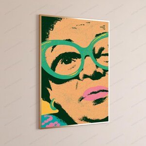 May include: A rectangular wall art print featuring a stylized, high-contrast portrait of a person wearing large, bright green cat-eye glasses. The artwork uses a bold color palette of peach, dark green, and pink, framed in a thin gold metal border.