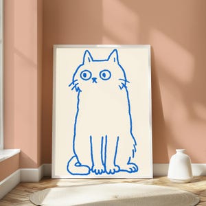 May include: A framed wall art print featuring a simple, hand-drawn blue line illustration of a cat with wide, circular eyes and a small nose, set against a solid cream-colored background within a thin, white rectangular frame.