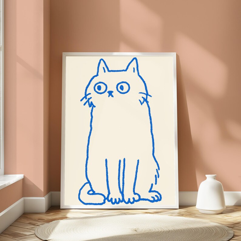 May include: A framed wall art print featuring a simple, hand-drawn blue line illustration of a cat with wide, circular eyes and a small nose, set against a solid cream-colored background within a thin, white rectangular frame.