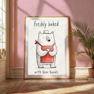 Puede incluir: Una l&aacute;mina enmarcada que muestra un oso blanco con un delantal rojo y un peque&ntilde;o gorro de chef. El oso est&aacute; de pie con las patas juntas. El texto "Freshly baked" aparece arriba y "with bear hands." est&aacute; impreso en la parte inferior.