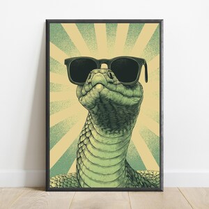 May include: A framed wall art print featuring a detailed illustration of a green snake wearing black sunglasses. The snake is positioned against a background of alternating light yellow and muted green sunburst rays, creating a graphic design.