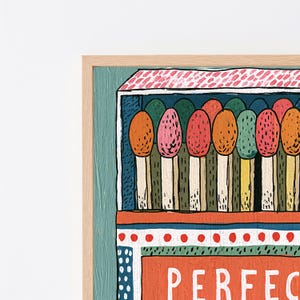 Op de afbeelding: Een ingelijste kunstdruk met een gestileerde illustratie van een luciferdoosje met kleurrijke luciferkoppen in tinten oranje, roze en groen. Het doosje heeft een rood-witte patroon bovenkant en een rode basis met de gedeeltelijke witte tekst PERFEC zichtbaar.