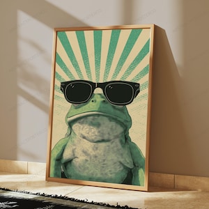 May include: A rectangular wall art print featuring a green frog wearing large black sunglasses. The background consists of a light beige and green sunburst pattern with a dotted texture. The frog is centered, facing forward with a neutral expression.