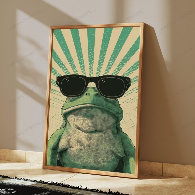 May include: A rectangular wall art print featuring a green frog wearing large black sunglasses. The background consists of a light beige and green sunburst pattern with a dotted texture. The frog is centered, facing forward with a neutral expression.