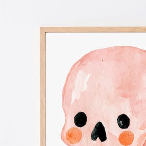 Puede incluir: Una ilustraci&oacute;n en acuarela enmarcada de un cr&aacute;neo de color rosa p&aacute;lido con grandes ojos circulares negros, una peque&ntilde;a nariz triangular negra y mejillas circulares de color naranja brillante sobre un fondo blanco en un marco de madera clara.