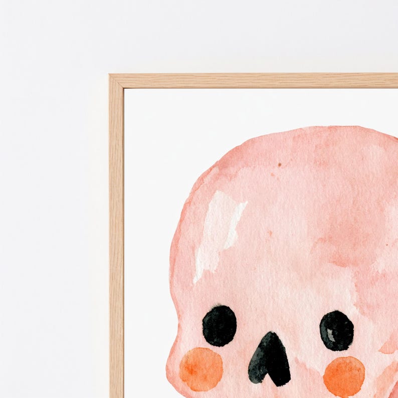 Puede incluir: Una ilustraci&oacute;n en acuarela enmarcada de un cr&aacute;neo de color rosa p&aacute;lido con grandes ojos circulares negros, una peque&ntilde;a nariz triangular negra y mejillas circulares de color naranja brillante sobre un fondo blanco en un marco de madera clara.