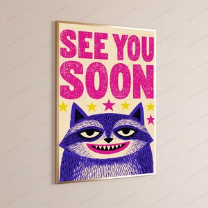 May include: A rectangular wall art print featuring the bold pink text SEE YOU SOON at the top. Below the text is a stylized purple raccoon face with a wide grin showing white teeth, set against a cream background with small yellow and pink stars.